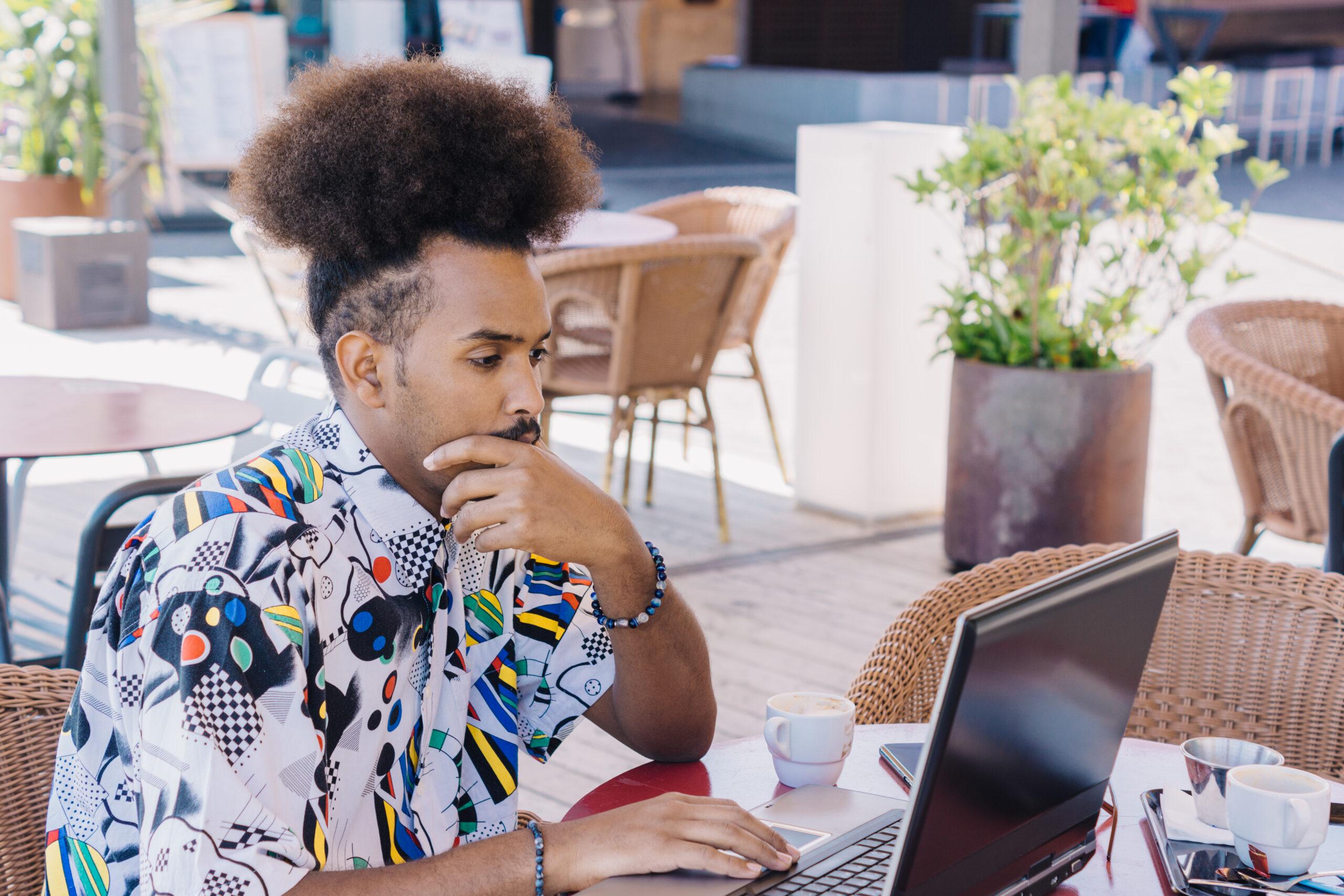 horizontal image of young black guy with afro hair tied back working with a laptop at the terrace table of a coffee shop